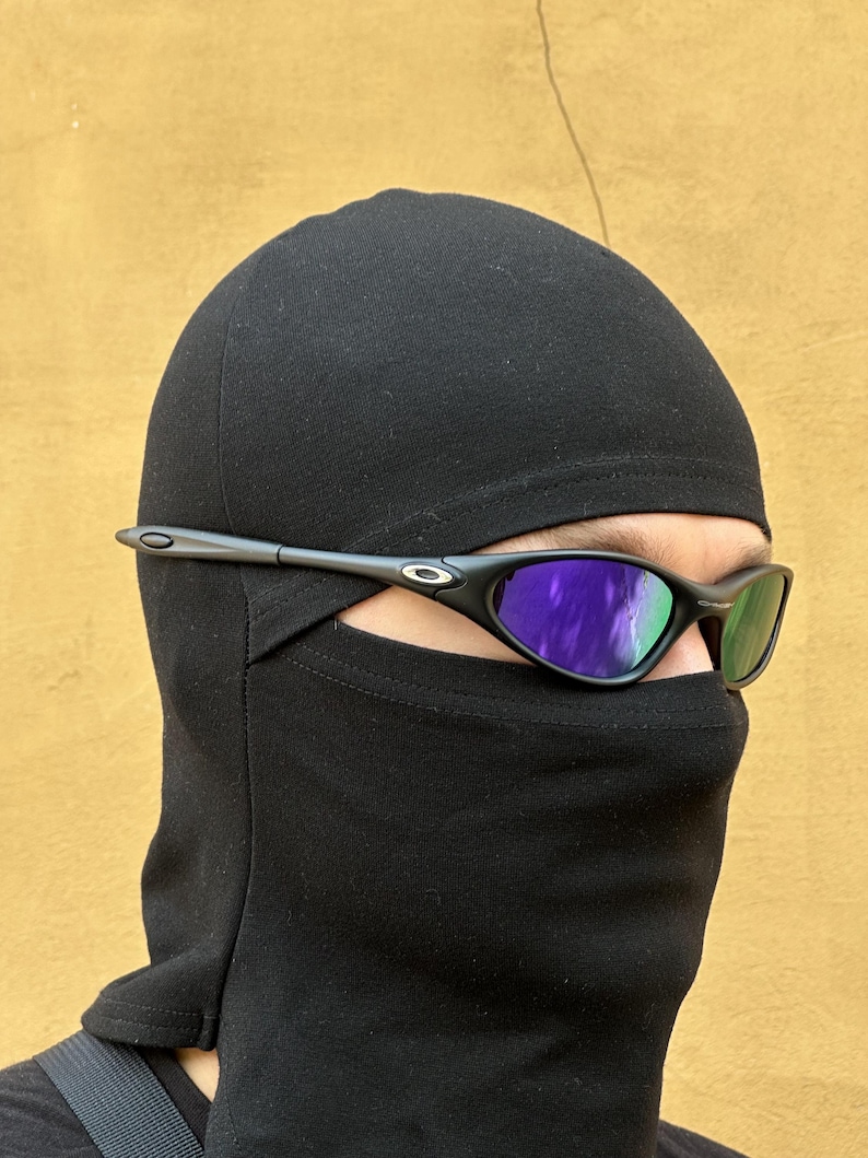May include: Black sunglasses with purple-tinted lenses are worn over a black balaclava. The sunglasses have a thin, dark frame. The background is a solid, light brown color.