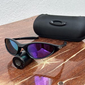 May include: Black sunglasses with purple lenses and a black case. The sunglasses are resting on a small, cylindrical object. The lenses reflect a purple hue, and the frame is black. The case is also black and has a subtle logo.