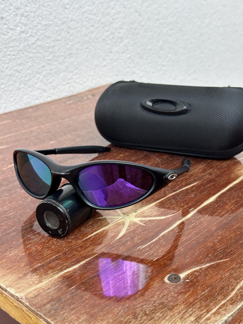 May include: Black sunglasses with purple lenses and a black case. The sunglasses are resting on a small, cylindrical object. The lenses reflect a purple hue, and the frame is black. The case is also black and has a subtle logo.