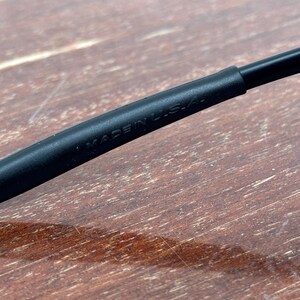 May include: Close-up of a black eyeglass frame arm with the text "MADE IN U.S.A." printed in white. The frame arm is made of a matte black material. The background is a wooden surface.