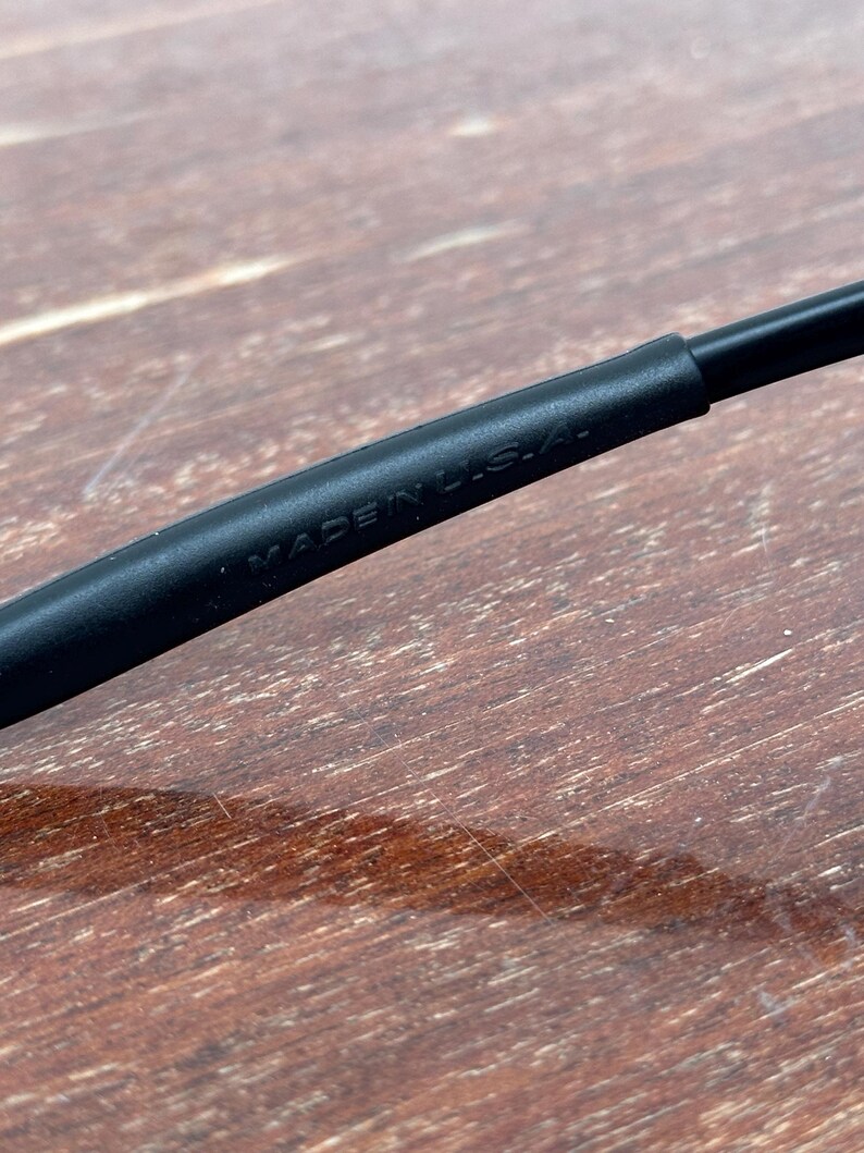 May include: Close-up of a black eyeglass frame arm with the text "MADE IN U.S.A." printed in white. The frame arm is made of a matte black material. The background is a wooden surface.