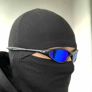 K&ouml;nnte beinhalten: Schwarze Sonnenbrille mit blau verspiegelten Gl&auml;sern. Die Sonnenbrille hat ein schlankes, modernes Design mit einem schwarzen Rahmen. Das Bild zeigt die Sonnenbrille in Kombination mit einer schwarzen Sturmhaube.