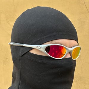 Puede incluir: Gafas de sol con montura plateada y lentes rojas y naranjas sobre un pasamontañas negro. Las gafas de sol tienen un diseño elegante y moderno. El pasamontañas cubre la cabeza y el cuello, dejando solo los ojos visibles.