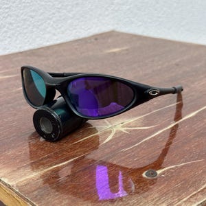 May include: Black sunglasses with purple lenses. The sunglasses have a sleek, curved design and are resting on a black cylindrical object. The lenses reflect a blue-green hue. The frame is black and has the brand logo on the side.