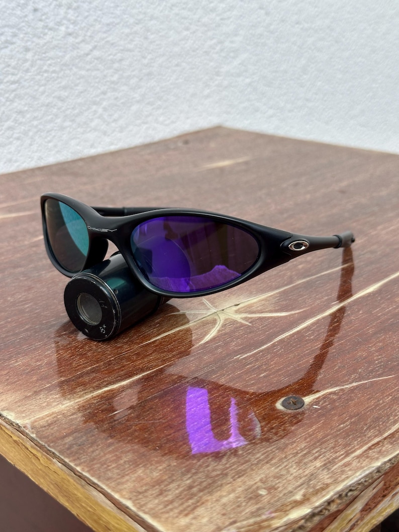 May include: Black sunglasses with purple lenses. The sunglasses have a sleek, curved design and are resting on a black cylindrical object. The lenses reflect a blue-green hue. The frame is black and has the brand logo on the side.