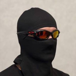 May include: A black balaclava with a neck gaiter, paired with black sunglasses featuring red and yellow lenses. The sunglasses have a black frame with a red accent. The balaclava covers the head and neck.
