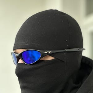 Puede incluir: Gafas de sol negras con lentes reflectantes azules se usan con un pasamontañas negro. Las gafas de sol tienen un diseño elegante y deportivo con montura negra. El pasamontañas cubre la parte inferior de la cara y la cabeza.