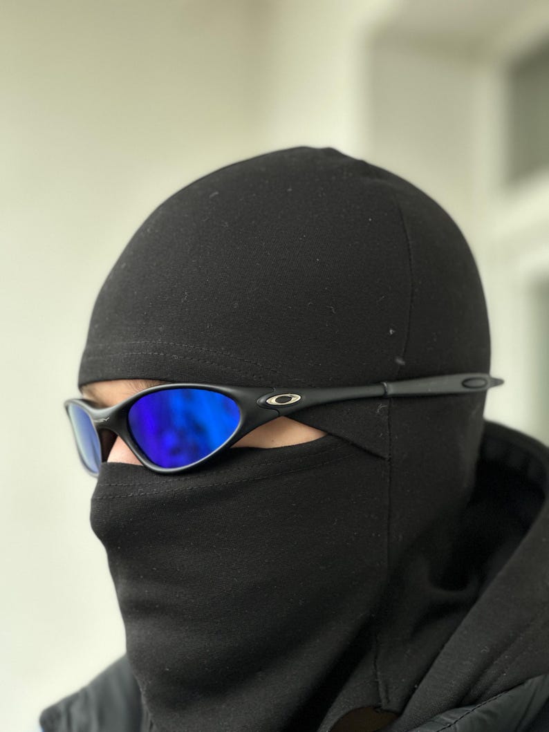 K&ouml;nnte beinhalten: Schwarze Sonnenbrille mit blau verspiegelten Gl&auml;sern wird mit einer schwarzen Sturmhaube getragen. Die Sonnenbrille hat ein schlankes, sportliches Design mit schwarzem Rahmen. Die Sturmhaube bedeckt das untere Gesicht und den Kopf.