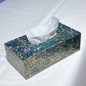 Puede incluir: Caja de pañuelos rectangular con un patrón único. La caja es principalmente turquesa con un diseño agrietado dorado y blanco. Los pañuelos blancos se sacan de la abertura superior. La caja está sobre una superficie blanca.