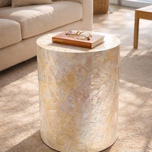 May include: A cylindrical side table with a mosaic shell design in cream, beige, and gold. A closed brown book and gold-rimmed glasses rest on top. The table is approximately 46 cm in diameter and 51 cm tall.