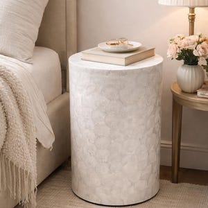 Könnte beinhalten: Ein zylindrischer Beistelltisch mit einer strukturierten, cremefarbenen Oberfläche. Auf dem Tisch liegen Bücher und ein kleiner weißer Teller mit einer Brille. Der Tisch ist etwa 50 cm hoch und steht auf einem neutralfarbenen Teppich.