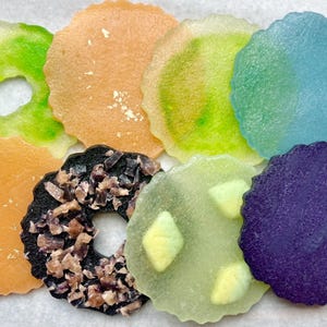 May include: A collection of colorful, circular candies in various hues, including green, orange, blue, and purple. Some candies have a hole in the center, while others are decorated with edible embellishments. The candies have a slightly translucent appearance.