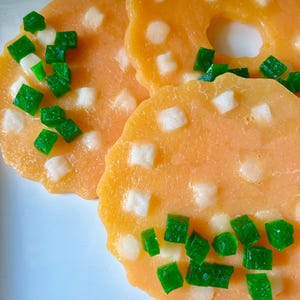 May include: Three round, scalloped-edge orange soap bars with a translucent texture. Each bar is embedded with small, square-shaped pieces of white and vibrant green material scattered across the surface, creating a textured, mosaic-like appearance.