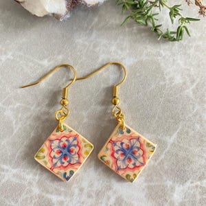 Könnte beinhalten: Ein Paar goldfarbene Hängeohrringe. Jeder Ohrring hat einen rautenförmigen Fliesen mit einem floralen Design in Blau, Rot und Grün. Die Fliesen sind cremefarben mit einem goldfarbenen Haken.