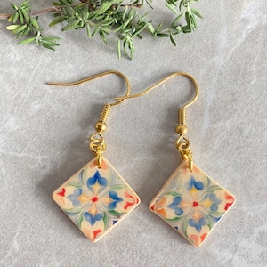 Può includere: Un paio di orecchini pendenti color oro. Ogni orecchino presenta una piastrella quadrata color crema con un motivo floreale blu, rosso e giallo. Gli orecchini sono esposti su una superficie grigio chiaro con rametti verdi.