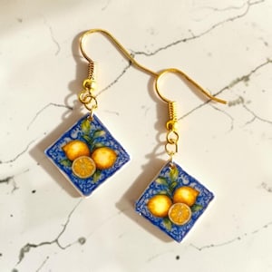 Può includere: Un paio di orecchini pendenti color oro. Ogni orecchino presenta un motivo a piastrella quadrata blu e bianca con un motivo a limone. I limoni sono gialli con foglie verdi e si vede anche una fetta di limone. Gli orecchini hanno una chiusura a gancio.