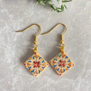 May include: A pair of gold-colored dangle earrings. Each earring features a square, cream-colored tile with a floral design in red, blue, orange, and yellow. The earrings have a hook closure.
