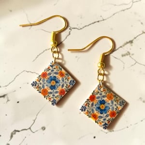 Op de afbeelding: Een paar goudkleurige oorhangers. Elke oorbel heeft een vierkante hanger met een bloemenpatroon in blauw, oranje en wit. De oorbellen hebben een haaksluiting.