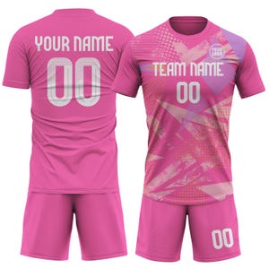 May include: A pink soccer uniform set, featuring a jersey and shorts. The jersey has "YOUR NAME" and the number "00" on the back, and "TEAM NAME" and "00" on the front. The design includes abstract patterns and a logo.