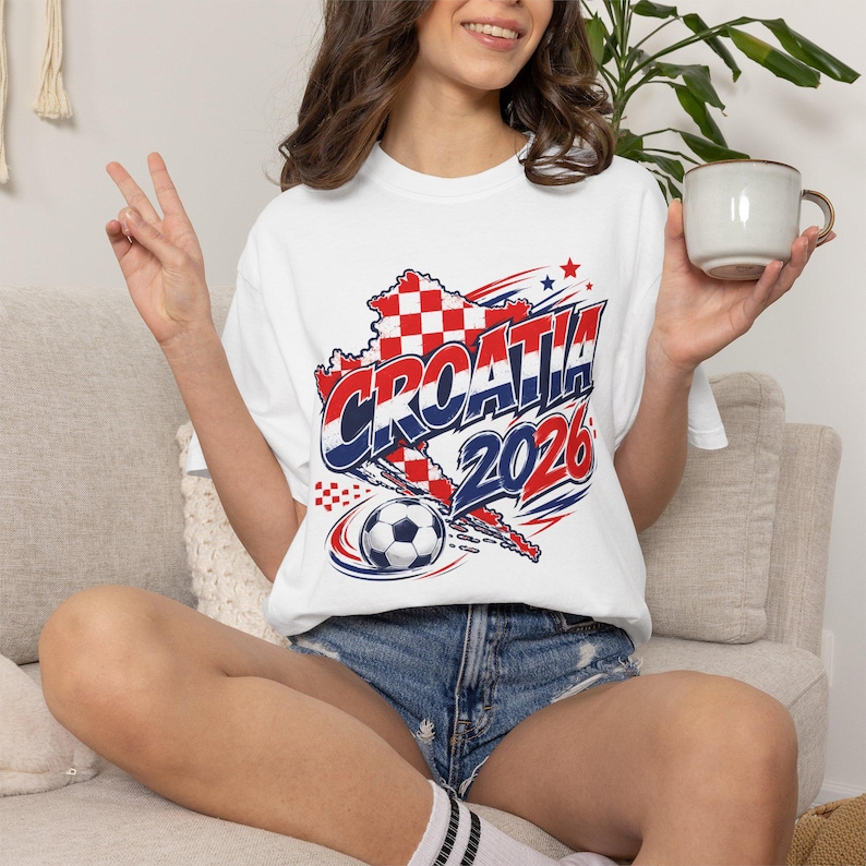 May include: White t-shirt with a graphic featuring the word "CROATIA" in red and blue, a map outline, a soccer ball, and the year "2026". The shirt is worn with denim shorts.