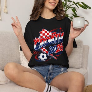 May include: Black t-shirt with a Croatia 2026 graphic. The design features the Croatian flag colors and a soccer ball. The shirt is a crew neck and is paired with denim shorts.