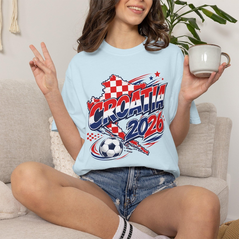 May include: Light blue t-shirt featuring a graphic of the Croatian flag, the word "CROATIA" in red and blue, and the year "2026". A soccer ball graphic is also included. The shirt is worn by a person.