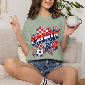 May include: A light green t-shirt with a graphic featuring the word "CROATIA" in blue and red, a map outline, a soccer ball, and the year "2026". The shirt is worn with denim shorts.