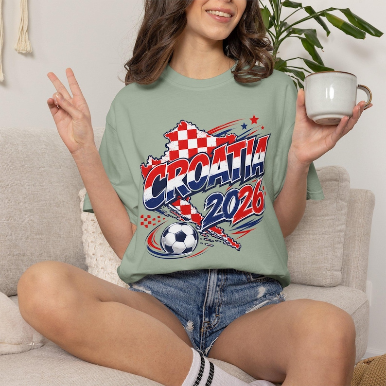 May include: A light green t-shirt with a graphic featuring the word "CROATIA" in blue and red, a map outline, a soccer ball, and the year "2026". The shirt is worn with denim shorts.