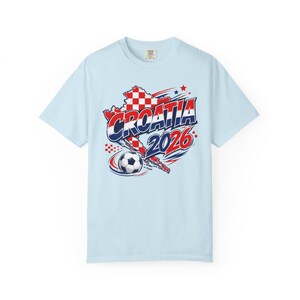 May include: Light blue t-shirt featuring a graphic design related to Croatia. The design includes the word "CROATIA" in red and white, the year "2026", a soccer ball, and a map outline of Croatia in red and white checkered pattern.