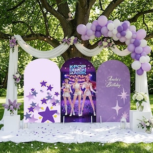 Pode incluir: Um cenário de festa com três painéis arqueados. O painel central exibe "K-POP DEMON HUNTERS" com figuras animadas. O painel esquerdo tem estrelas roxas, e o direito diz "Amelia's Birthday". Um arco de balões e arranjos florais decoram a cena.