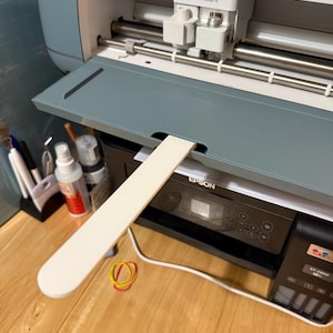 May include: A Cricut Maker 4 machine in a light blue-gray color, with the lid open. A white, rectangular piece of material is being fed into the machine. An Epson printer is below the Cricut machine.