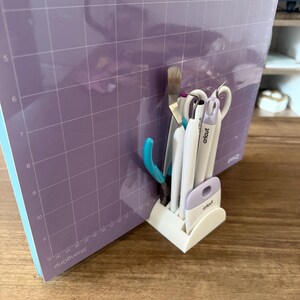 Puede incluir: Un organizador de herramientas Cricut blanco que contiene varias herramientas de manualidades, incluyendo tijeras, una espátula y pinzas. Las herramientas están colocadas frente a una alfombrilla de corte morada grande con un patrón de cuadrícula. La alfombrilla está etiquetada como "StrongGrip".