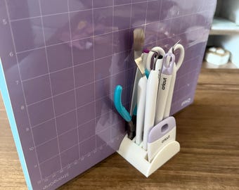 May include: A white Cricut tool organizer holding various crafting tools, including scissors, a spatula, and tweezers. The tools are positioned in front of a large, purple cutting mat with a grid pattern. The mat is labeled "StrongGrip".