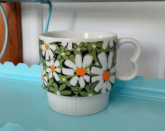 May include: White ceramic mug with a daisy pattern in green, black, and orange. The mug has a heart-shaped handle and a ridged base. The mug is sitting on a light blue surface.
