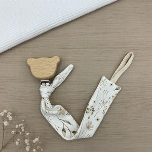 Könnte beinhalten: Ein Schnullerhalter aus Holz in Bärenform mit einem Stoffband mit floralem Muster. Der Riemen hat einen cremefarbenen Hintergrund mit braunen und beigen floralen Designs. Der Clip ist aus Naturholz und hat einen Metallverschluss.