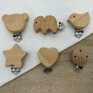 Könnte beinhalten: Eine Sammlung von sechs Holzclips für Babyschnuller in verschiedenen Formen: ein Bär, ein Elefant, ein Vogel, ein Stern, ein Herz und eine runde Form mit Löchern. Jeder Clip hat einen silberfarbenen Metallverschluss. Die Clips sind aus hellem Holz.
