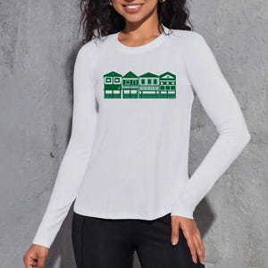 May include: White long-sleeved top featuring a green graphic of a row of buildings. The buildings have different architectural styles. The top is made of a soft, lightweight fabric.
