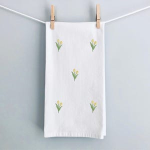 May include: A white hand towel with embroidered yellow tulips and green leaves, hanging from a clothesline with wooden clothespins. The towel is rectangular and has a simple, clean design, perfect for a bathroom or kitchen.