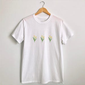 May include: White t-shirt with three embroidered yellow tulip flowers with green stems and leaves. The crew neck tee shirt is hanging on a wooden hanger.