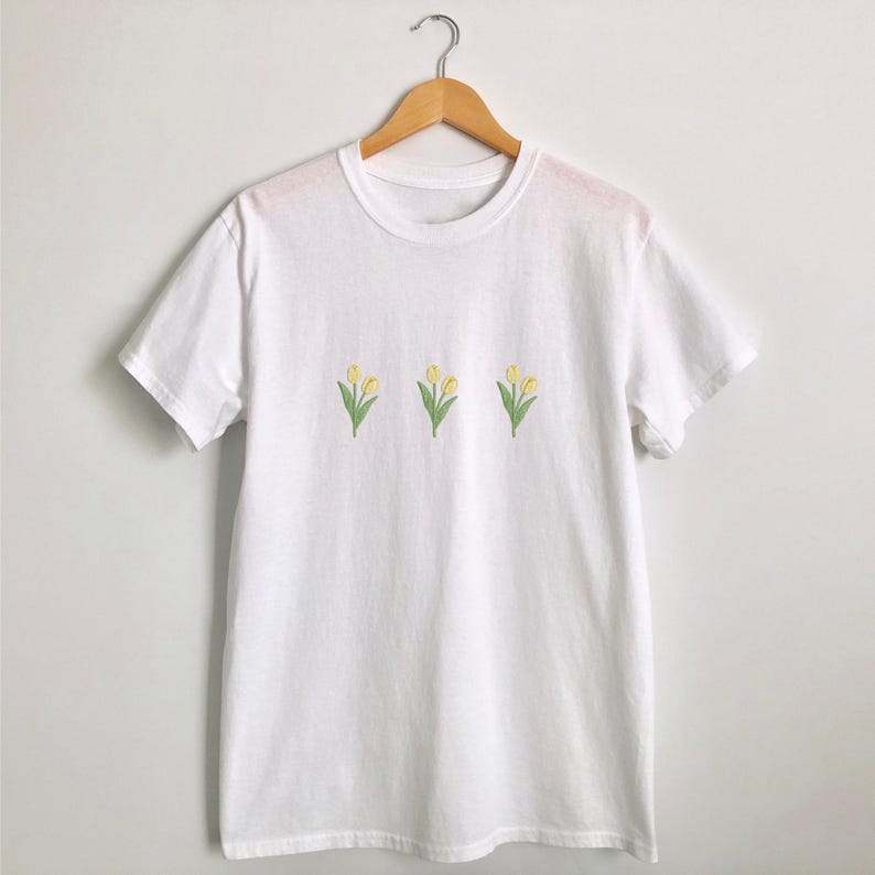May include: White t-shirt with three embroidered yellow tulip flowers with green stems and leaves. The crew neck tee shirt is hanging on a wooden hanger.