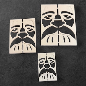May include: Three off-white, rectangular face-shaped puzzle pieces with a mustache and beard design. The pieces are arranged on a dark surface. The faces have a stylized, cutout design.