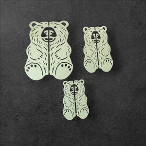 May include: Three light-colored bear-shaped cutouts with detailed etched designs. The bears are in different sizes, with the largest at the top and the smallest at the bottom. The bears have a split down the middle.