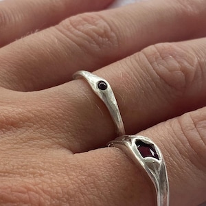 May include: Two silver rings with a hammered finish. One ring has a small, round, dark red gemstone, the other a larger, square-cut, dark red gemstone. The rings are shown on a finger.