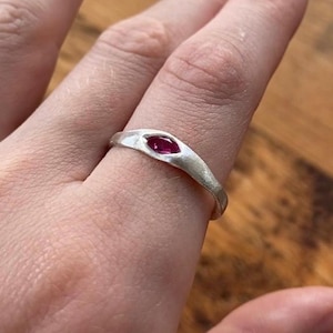 Könnte beinhalten: Ein silberner Ring mit einem marquiseförmigen, tiefroten Edelstein. Der Ring hat ein schlichtes, elegantes Design mit leicht strukturierter Oberfläche. Der Edelstein ist in einer Lünettenfassung gefasst, und das Band ist glatt und abgerundet.