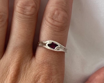 May include: A silver ring with a textured band and a dark red, diamond-shaped gemstone. The ring is worn on a finger, showcasing the intricate details of the metalwork and the vibrant color of the stone. The design is organic and elegant.