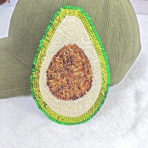 Pode incluir: Boné de beisebol verde-oliva com um design de abacate com lantejoulas. O abacate apresenta um caroço castanho, polpa branca e casca verde. O boné é feito de tecido de veludo cotelê.