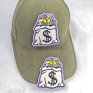 Puede incluir: Gorra de béisbol verde oliva con textura de pana. La gorra presenta dos parches bordados de una bolsa de dinero con un signo de dólar y lingotes de oro. Los parches tienen un borde morado.
