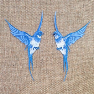 Könnte beinhalten: Zwei gestickte Aufnäher mit blau-weißen Vögeln mit ausgebreiteten Flügeln. Die Vögel haben lange, fließende Schwanzfedern und sind vor einem neutralen, strukturierten Hintergrund platziert. Diese Aufnäher können zum Verzieren von Kleidung oder Accessoires verwendet werden.