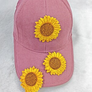 Könnte beinhalten: Staubrosa Baseballkappe mit drei gestickten gelben Sonnenblumen. Die Kappe hat einen gebogenen Schirm und einen Stoffverschluss. Die Sonnenblumen haben detaillierte gelbe Blütenblätter und braune Zentren.