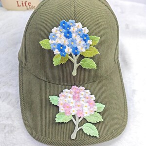 以下が含まれることがあります： オリーブグリーンのコーデュロイベースボールキャップ。刺繍されたアジサイの花が特徴です。キャップの上部には青と白のアジサイ、つばにはピンクと白のアジサイがあります。花には緑の葉と茎があります。